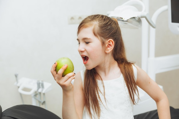 The girl at the reception at the pediatric dentist smiles and laughs a little girl holds a big green apple in her hands children health concept