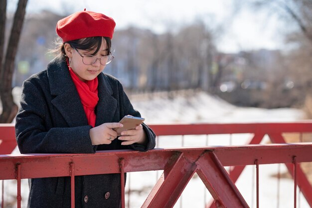 a girl in a red beret and scarf is standing on the bridge with a phone in her hands