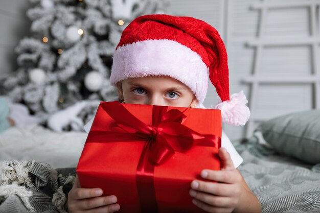 Girl in a red cap with a Christmas present. She lies on the bed, hugs the box against the background of the Christmas tree.