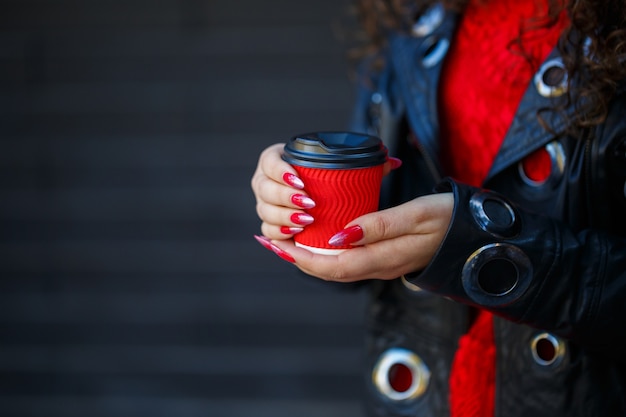 Girl in a red dress and a black jacket drinks coffee from a red paper cup