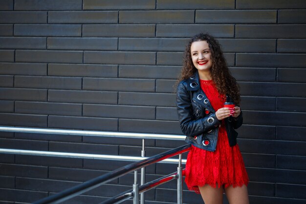 Girl in a red dress and a black jacket drinks coffee from a red paper cup