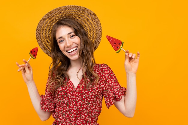 Girl in a red dress and hat on a yellow background with a wide smile and lollipops in her hands