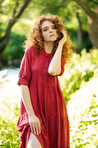 Girl in red dress posing by the river lady with curly hair\
standing by a tree vacation portrait