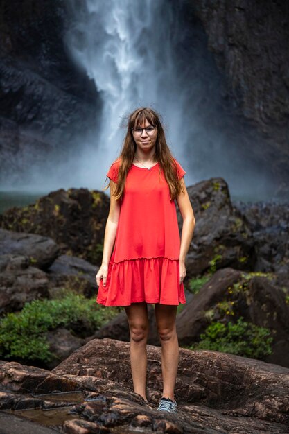 girl in red dress stands under huge waterfall, dramatic view of wallaman falls, queensland, aus