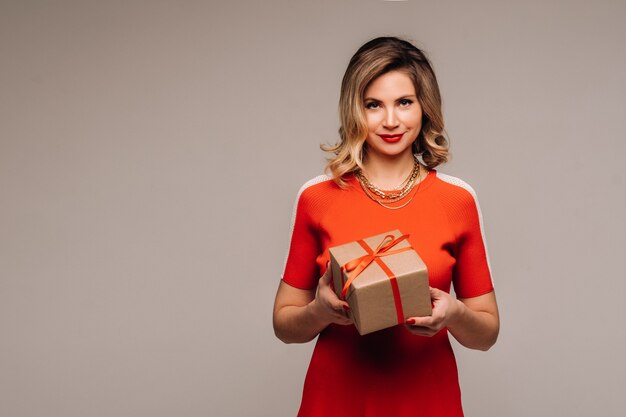 A girl in a red dress stands with gifts in her hands on a gray wall.