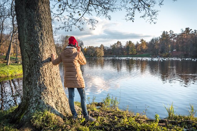 ヘッドホンをつけた赤い帽子をかぶった女の子が秋の公園の景色を楽しんでいます後ろからの眺め