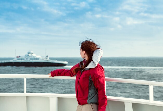 The girl in a red jacket on the ferryboat looks towards the sea, where the liner floats