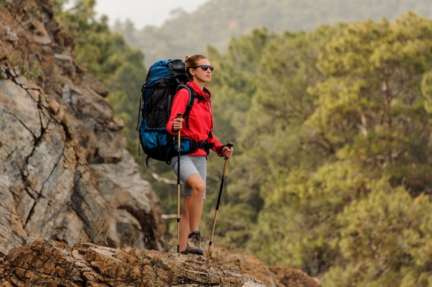 Girl in red jacket standing on the rock with hiking backpack