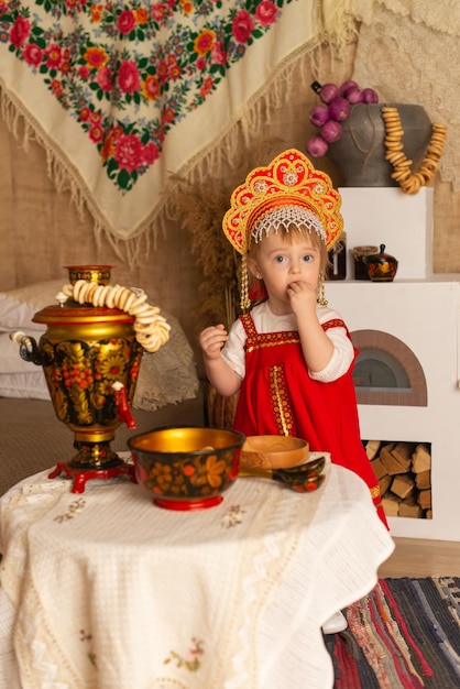 サモワールとハンドルを持つテーブルの近くの赤い民族衣装の女の子パンケーキの日
