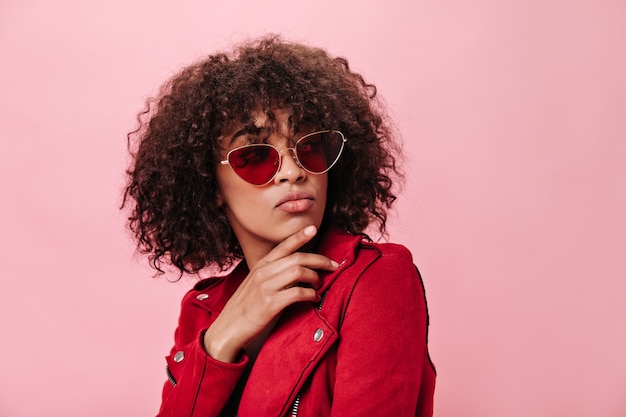 Girl in red outfit and sunglasses posing thoughtfully on pink wall