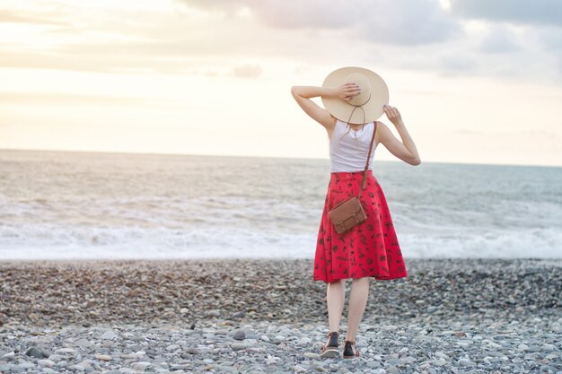 Girl in a red skirt and hat is standing by the sea, sunset. View from the back