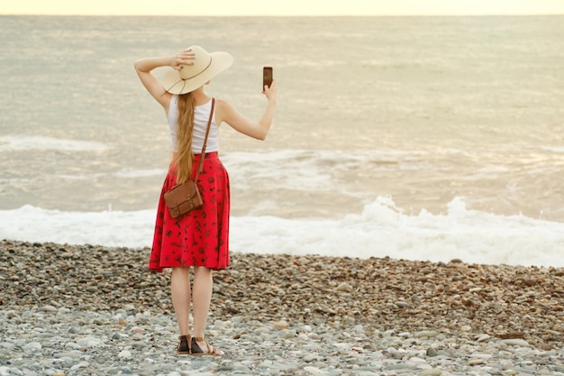 Girl in red skirt and hat making selfie on the beach