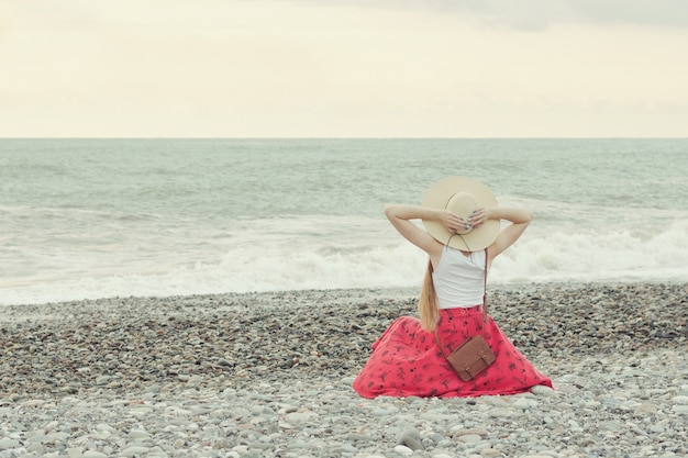 Girl in red skirt and hat sits on the seashore. Sunset time. Back view