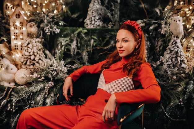 A girl in a red suit is sitting on a chair near the Christmas background. a woman in red on the background of Christmas trees and small Christmas houses