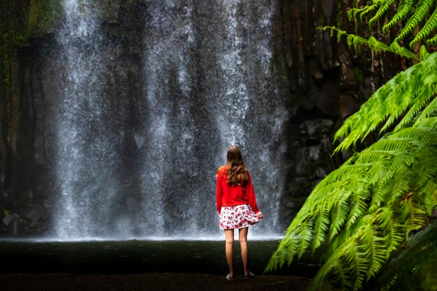 A girl in a red sweatshirt and red and white skirt stands over a powerful tropical waterfall in aus