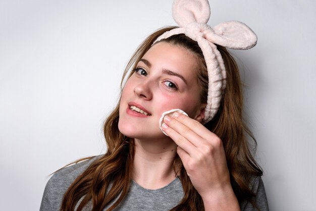 Girl removing makeup from her face with cotton pad on gray