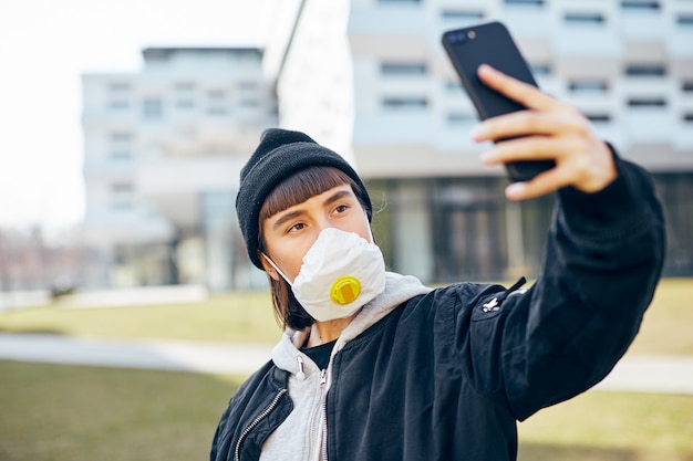 Girl in respiratory protection mask making selfies on the street, millenial female in casual wear and black hat doing self photos on her phone outdoor in masks