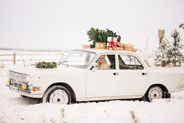 The girl rides in a retro car decorated with a Christmas tree and presents in a snowy forest. The concept of a winter Christmas photo shoot