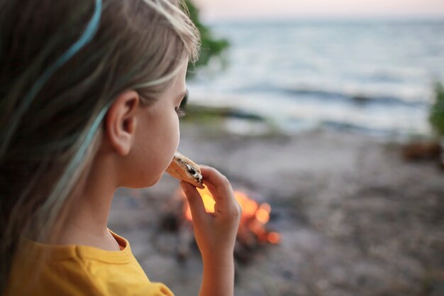 キャンプの伝統的な旅行の食べ物の間に火の炎の上でスモアを作るためにマシュマロを焼く女の子