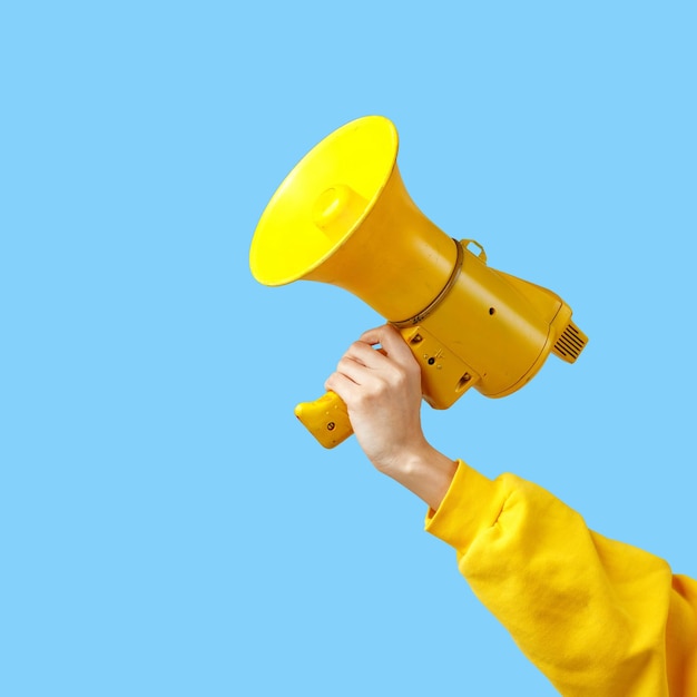 A girl's hand in yellow clothes holds a yellow megaphone on a blue background