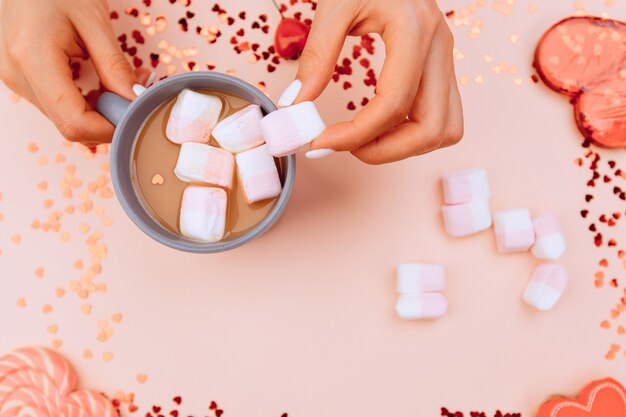Foto girl's handen zetten marshmallows in een kopje koffie en op trendy roze papier. valentijnsdag en vrouwendag concept. bovenaanzicht