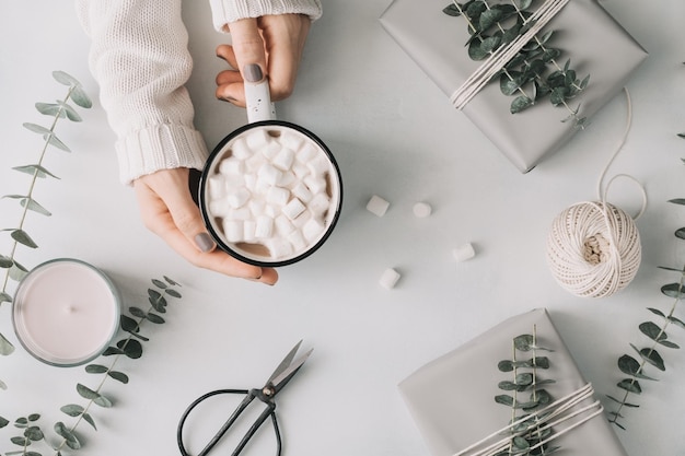 Le mani della ragazza tengono una tazza con cioccolata calda e marshmallow tra scatole regalo ed eucalipto su un tavolo bianco il concetto di inverno e regali di confezionamento vista dall'alto piatta stile minimal