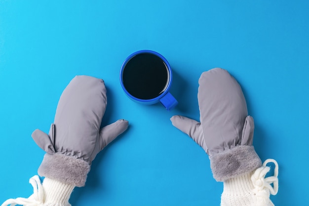 A girl's hands in mittens and a cup of black coffee on a blue background. Hot drink and mittens.