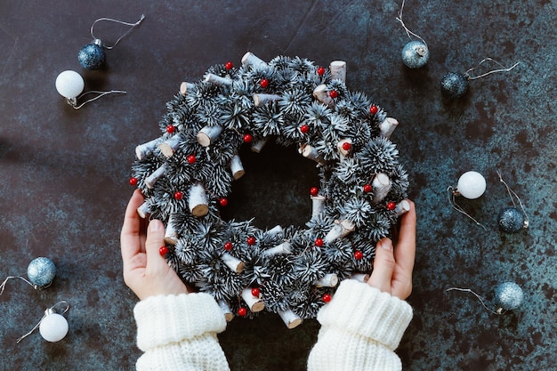 Girl's hands in a winter white sweater hold a Christmas holiday wreath on a blue table among by New Year's decor Top view flat lay