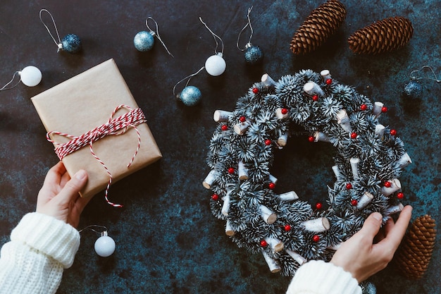 Girl's hands in a winter white sweater tie New Year's gift box in craft paper on a table among Christmas decor Top view flat lay