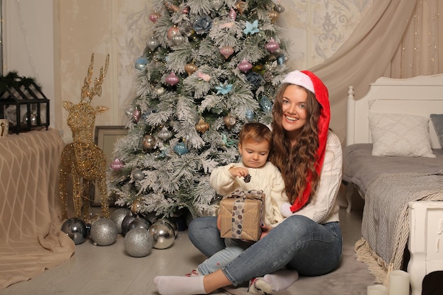 a girl in a Santa Claus hat at the Christmas tree next to her daughter with a gift box