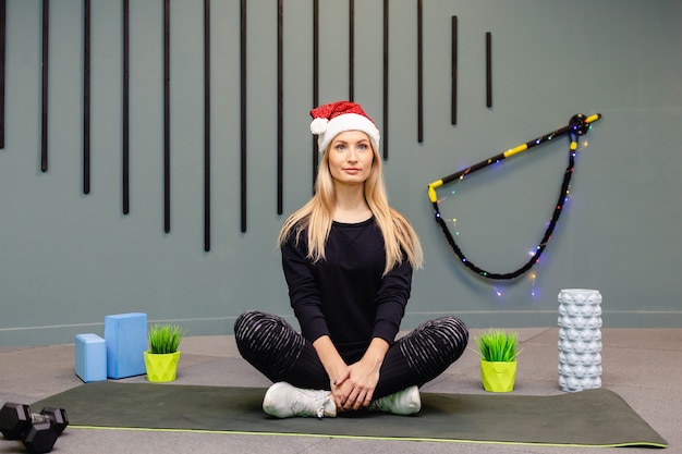 Девушка в шляпе Санта-Клауса делает фитнес-упражнения в тренажерном зале, в черной спортивной форме