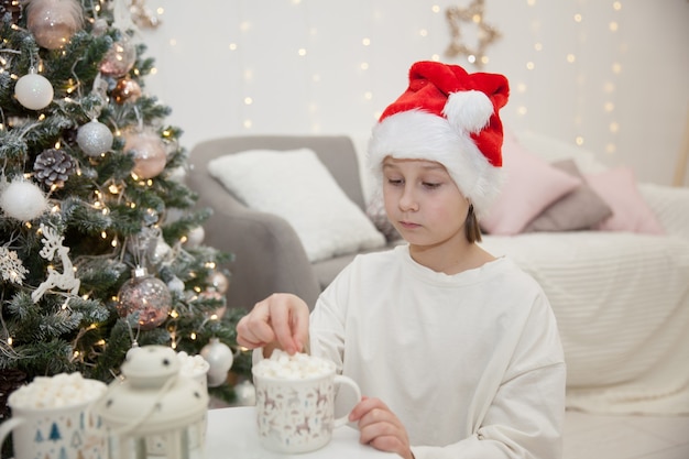 Girl in Santa Claus hat drinks cocoa with marshmallows amid Christmas tree