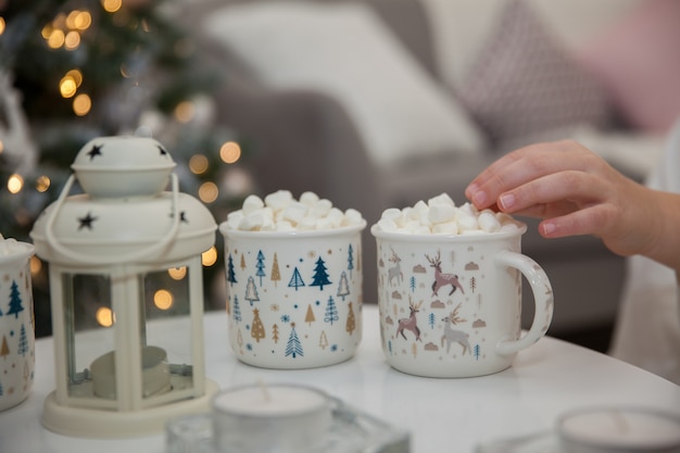 Girl in Santa Claus hat drinks cocoa with marshmallows amid Christmas tree