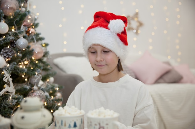 Girl in Santa Claus hat drinks cocoa with marshmallows amid Christmas tree