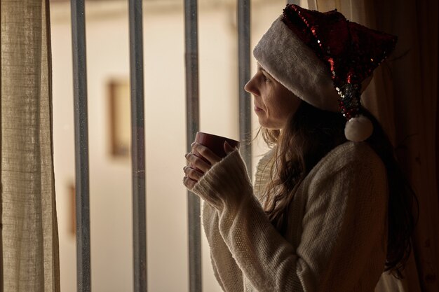 Girl in santa claus hat having a cup of coffee and looking out the window