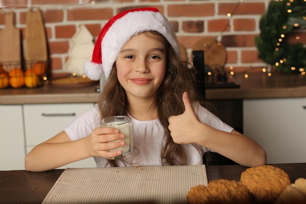 A girl in a Santa Claus hat in the kitchen holds a glass of milk in her hands and shows clas