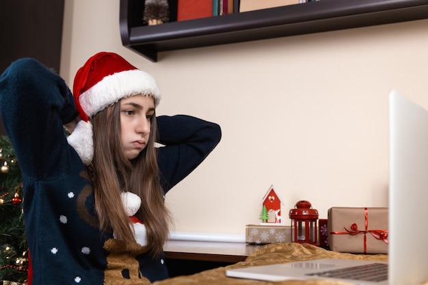 girl in santa claus hat talks using laptop for video call friends and parents. The room is festively decorated. Christmas during coronavirus. fatigue