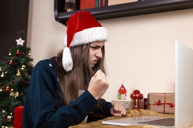 girl in santa claus hat talks using laptop for video call friends and parents. The room is festively decorated.