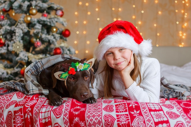 A girl in a Santa Claus hat with a dog lie on the bed near the Christmas tree