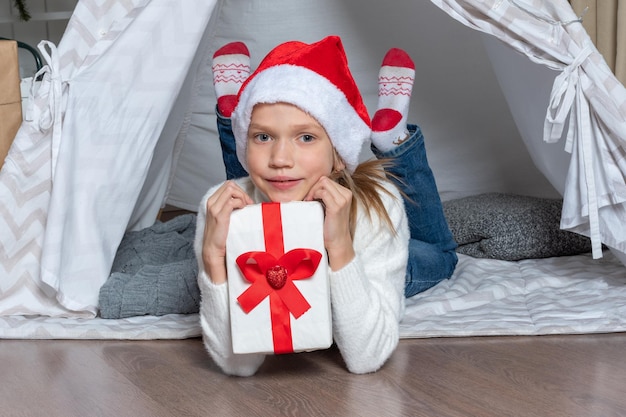 A girl in a Santa hat holds his big gift box for Christmas New Year lying in a children's tent wigwam in the nurser