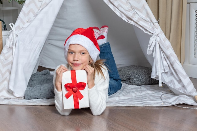 A girl in a santa hat holds his big gift box for christmas new\
year lying in a children\'s tent wigwam in the nurser