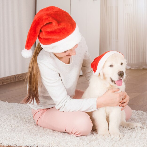 サンタの帽子をかぶった女の子は、サンタの帽子でレトリーバーの子犬を抱き締めて座っています