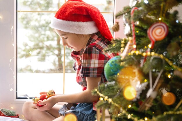 Girl in a Santa hat sits on the windowsill of house near the Christmas tree and holding glass balls