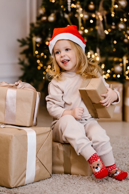 クリスマスツリーの背景にクリスマスプレゼントとサンタ帽子の女の子。自宅でクリスマスプレゼントを持った子供。