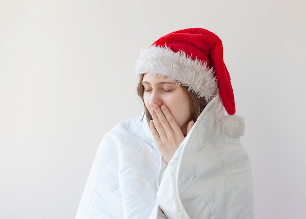 Девушка в новогодней шапке закуталась в белое одеяло и, зевнув, прикрыла рот ладонью ...