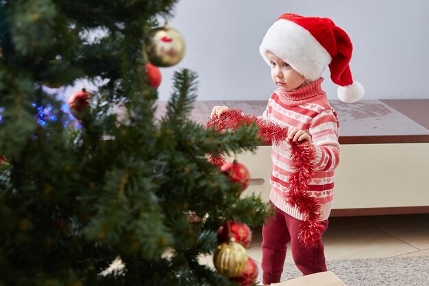 Girl in Santa's New Year's hat decorates the Christmas tree at home.