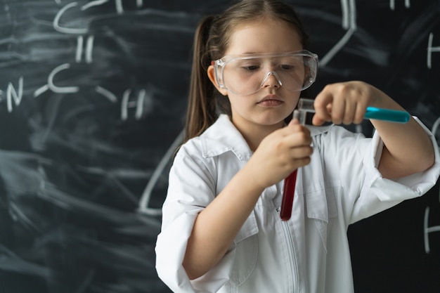 The girl schoolgirl is dressed in a white robe and goggles conduct an experiment in a chemistry less...