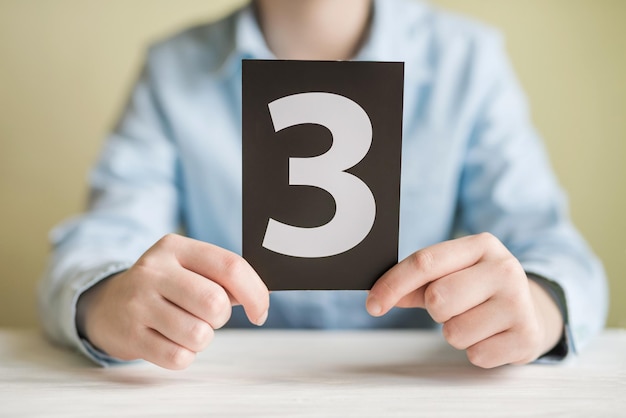 Photo a girl in a shirt holds a sign with the number 3 sitting at the table