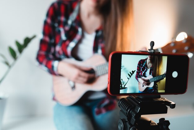 Foto ragazza in camicia e jeans scrive un vlog al telefono sul gioco delle ukulele.