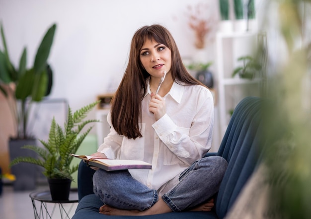 A girl in a shirt sits on a sofa in an apartment and writes something in a diary